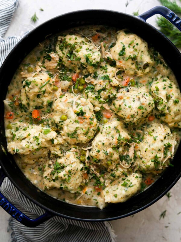 Finished spring chicken and dumplings fill a dark blue Staub cocotte. The cocotte sits atop a creamy white textured surface and is surrounded by a blue and white striped linen napkin and fresh herbs.