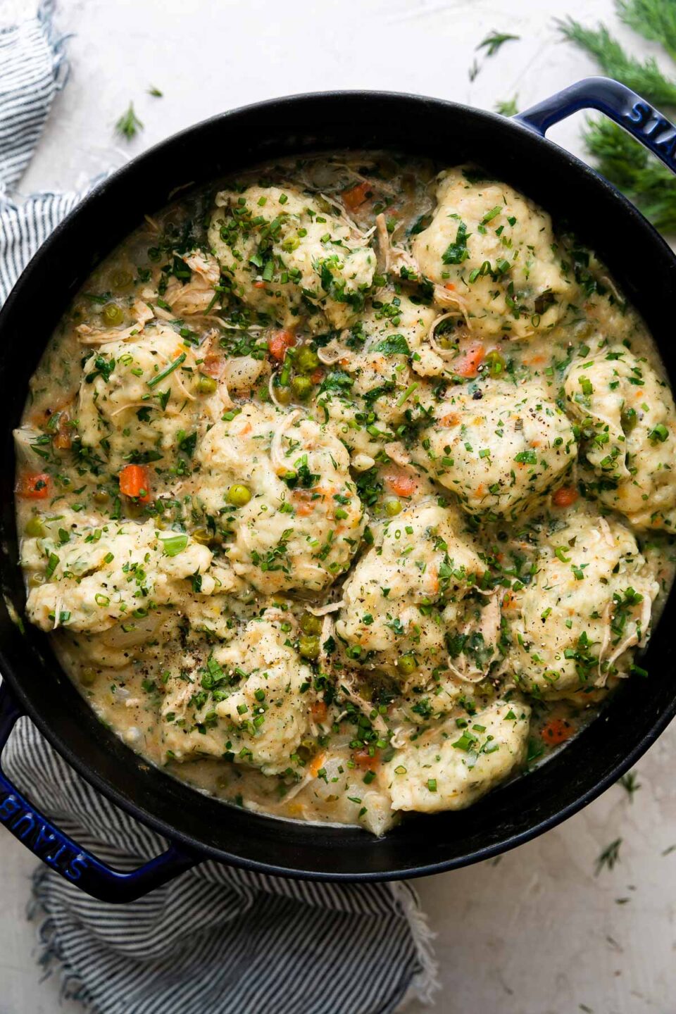 Finished spring chicken and dumplings fill a dark blue Staub cocotte. The cocotte sits atop a creamy white textured surface and is surrounded by a blue and white striped linen napkin and fresh herbs.