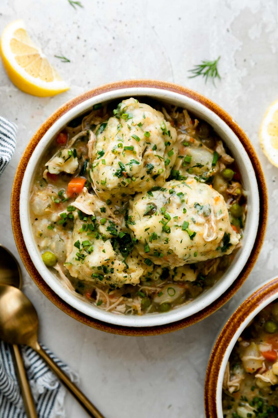 Two servings of homemade chicken and dumplings fill two brown rimmed ceramic bowls. The bowls sit atop a creamy white textured surface surrounded by lemon wedges, fresh herbs, a blue and white striped linen napkin and two gold spoons.