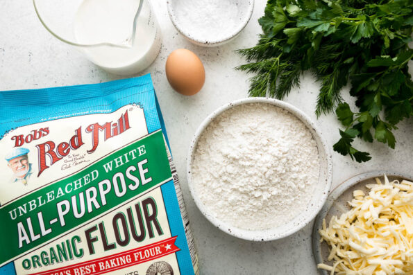 Spring chicken and dumplings drop dumpling ingredients arranged on a creamy white surface: Bob's Red Mill Organic All-Purpose Flour, baking powder, fresh herbs, eggs, cold unsalted butter, half & half.