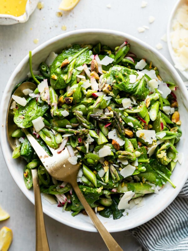 An overhead shot of finished Spring Vegetable Salad tossed in a Lemon Dijon Vinaigrette and finished with shaved parmesan and lemon zest inside of a large white ceramic bowl. A gold spoon and fork rest inside of the bowl for serving. The bowl sits atop a white textured surface with a blue and white striped linen napkin rest alongside the bowl and a small white speckled ceramic bowl filled with lemon dijon vinaigrette, loose lemon wedges, and small white plate with shaved parmesan surrounding the bowl of salad at center.