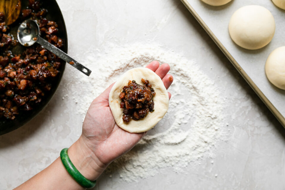 How to make char siu manapua, step 4: Assemble the manapua. A woman's hand holds a flattened piece of dough (roughly 3 1/2 inches in diameter) above a creamy white textured surface dusted with flour. Char siu filling has been scooped atop the flattened dough. To the left of the char siu topped dough ball is a skillet filled with char siu filling with a metal tablespoon resting inside and to the right of the flattened dough ball is a parchment lined baking sheet of proofed & risen dough balls.