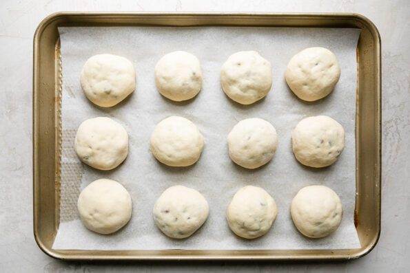 Char siu manapua filled bao buns are arranged on a parchment lined baking sheet for proofing. The baking sheet sits atop a creamy white textured surface.
