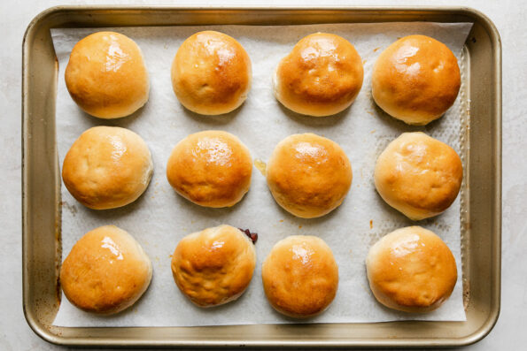 How to make manapua, step 6: Bake the manapua. Several baked char siu manapua buns are arranged on a parchment lined baking sheet. The baking sheet sits atop a creamy white textured surface.