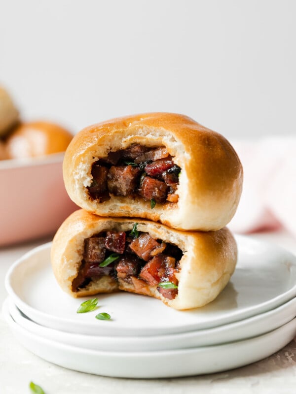 Three white plates topped with two baked manapua rest atop a creamy white textured surface. One of the baked manapua has been bitten into revealing the inside char siu filling. The plates are surrounded by a pink serving dish filled with more baked manapua, a light pink linen napkin, and loose thinly sliced green onion.