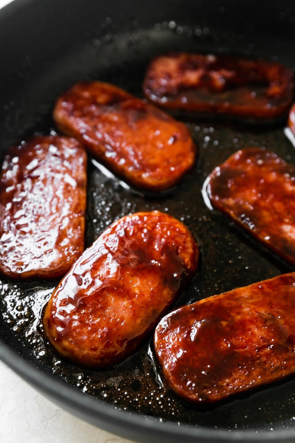 Spam glazed with teriyaki sauce pan fries in a large black skillet. The pan sits atop a creamy white textured surface.
