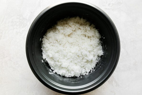 How to make spam musubi, step 1: Cook the rice. Cooked rice rests in the bottom of a pot-style rice cooker. The rice cooker rests atop a creamy white textured surface.