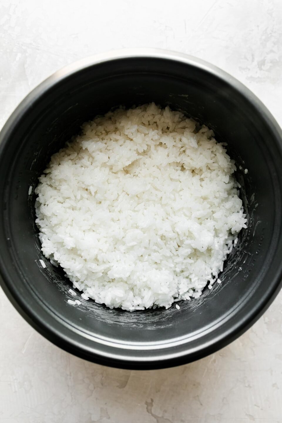 Cooked rice rests in the bottom of a pot-style rice cooker. The rice cooker rests atop a creamy white textured surface.