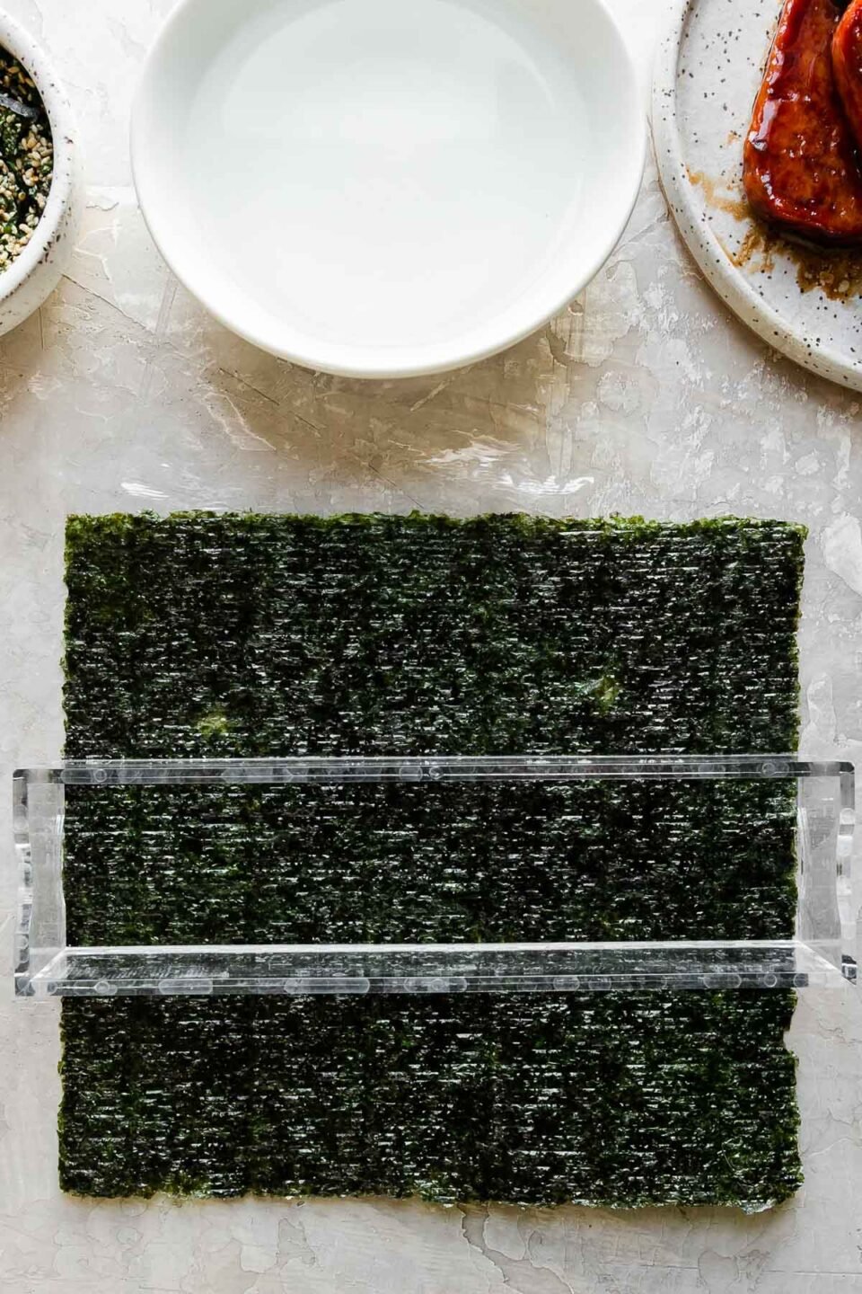 A close up of a single sheet of sushi nori rests atop a piece of plastic wrap that sits atop a creamy white textured surface. The outer box of a double musubi mold is arranged in the center of the sushi nori with the long edge of the mold running parallel with the long edge of the nori. Resting above the nori & the musubi mold is a small bowl filled with furikake seasoning with a spoon resting inside, another small white bowl filled with water, and a small speckled ceramic plate with pan-fried teriyaki Spam.