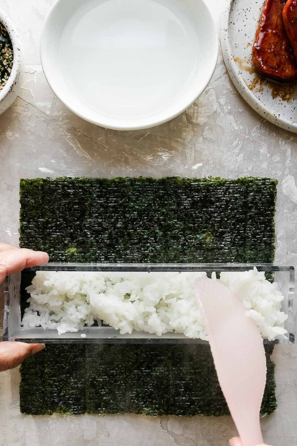 A close up of a woman's hands work to fill the outer box of a double musubi mold that rests atop a single sheet of sushi nori arranged on a piece of plastic wrap atop a creamy white textured surface. The woman stablizes the musubi mold with one hand while she uses a rice paddle in the other to gently add a layer of cooked rice to the inside of the mold. Resting above the nori & the musubi mold is a small bowl filled with furikake seasoning with a spoon resting inside, another small white bowl filled with water, and a small speckled ceramic plate with pan-fried teriyaki Spam.