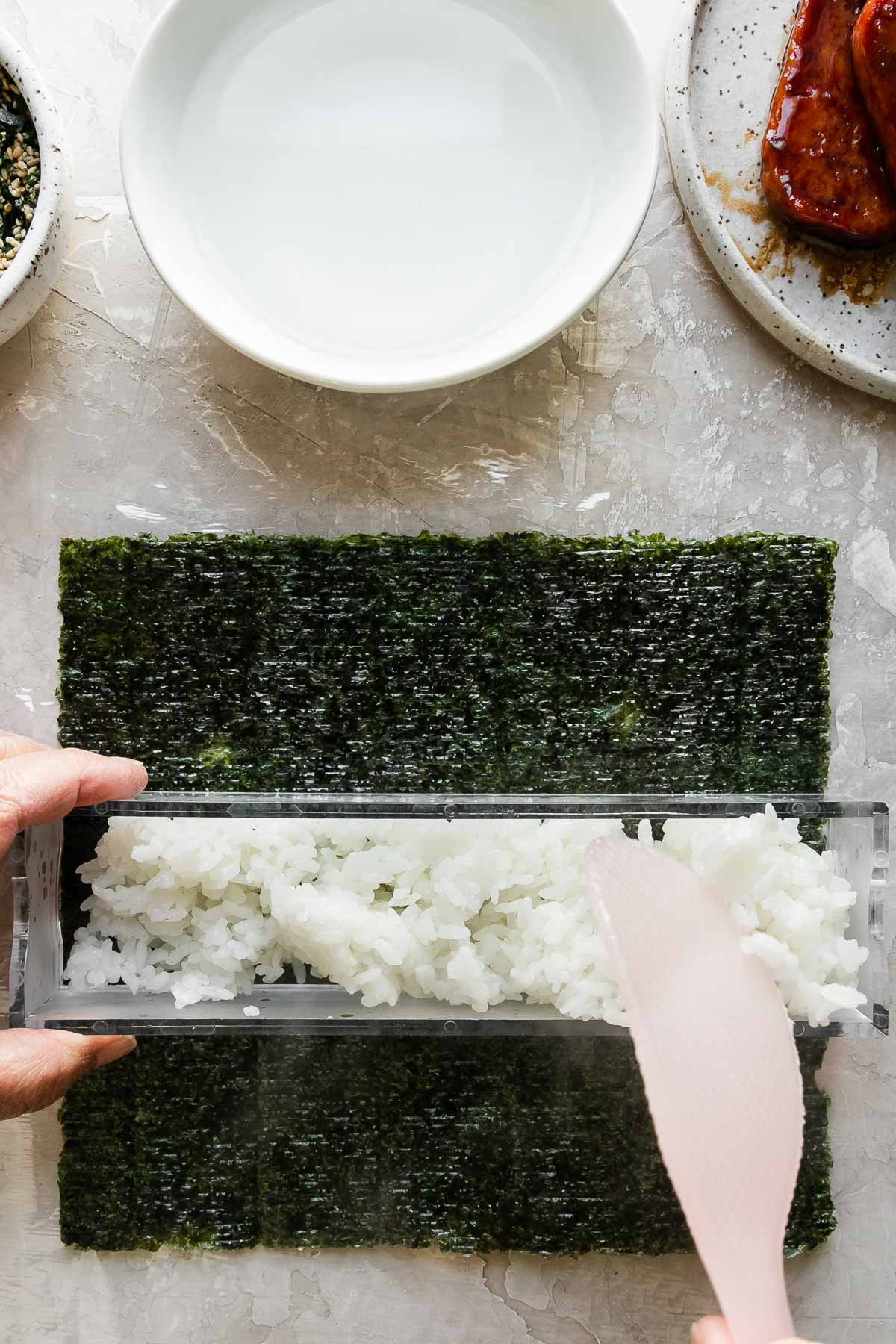 How to make Hawaiian spam musubi, step 5: Build the spam musubi. A close up of a woman's hands work to fill the outer box of a double musubi mold that rests atop a single sheet of sushi nori arranged on a piece of plastic wrap atop a creamy white textured surface. The woman stablizes the musubi mold with one hand while she uses a rice paddle in the other to gently add a layer of cooked rice to the inside of the mold. Resting above the nori & the musubi mold is a small bowl filled with furikake seasoning with a spoon resting inside, another small white bowl filled with water, and a small speckled ceramic plate with pan-fried teriyaki Spam.