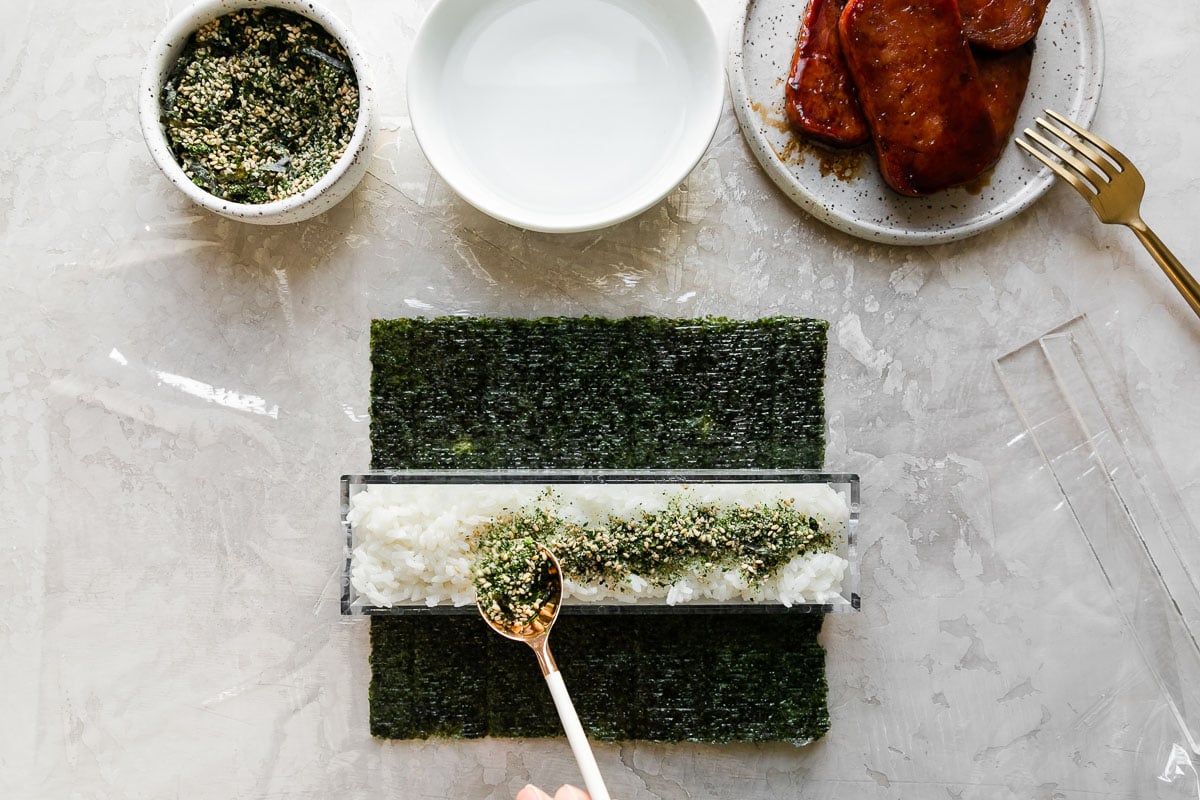 How to make spam musubi, step 5: Build the spam musubi. A woman's hand holds a white and gold spoon to sprinkle a generous amount of furikake seasoning atop a rice filled outer box of a double musubi mold. The musubi mold rests atop a single sheet of sushi nori arranged on a piece of plastic wrap atop a creamy white textured surface. Resting above the nori & the musubi mold is a small bowl filled with furikake seasoning with a spoon resting inside, another small white bowl filled with water, and a small speckled ceramic plate with pan-fried teriyaki Spam resting atop with a gold fork resting on the plate. The musubi mold lids rests alongside.
