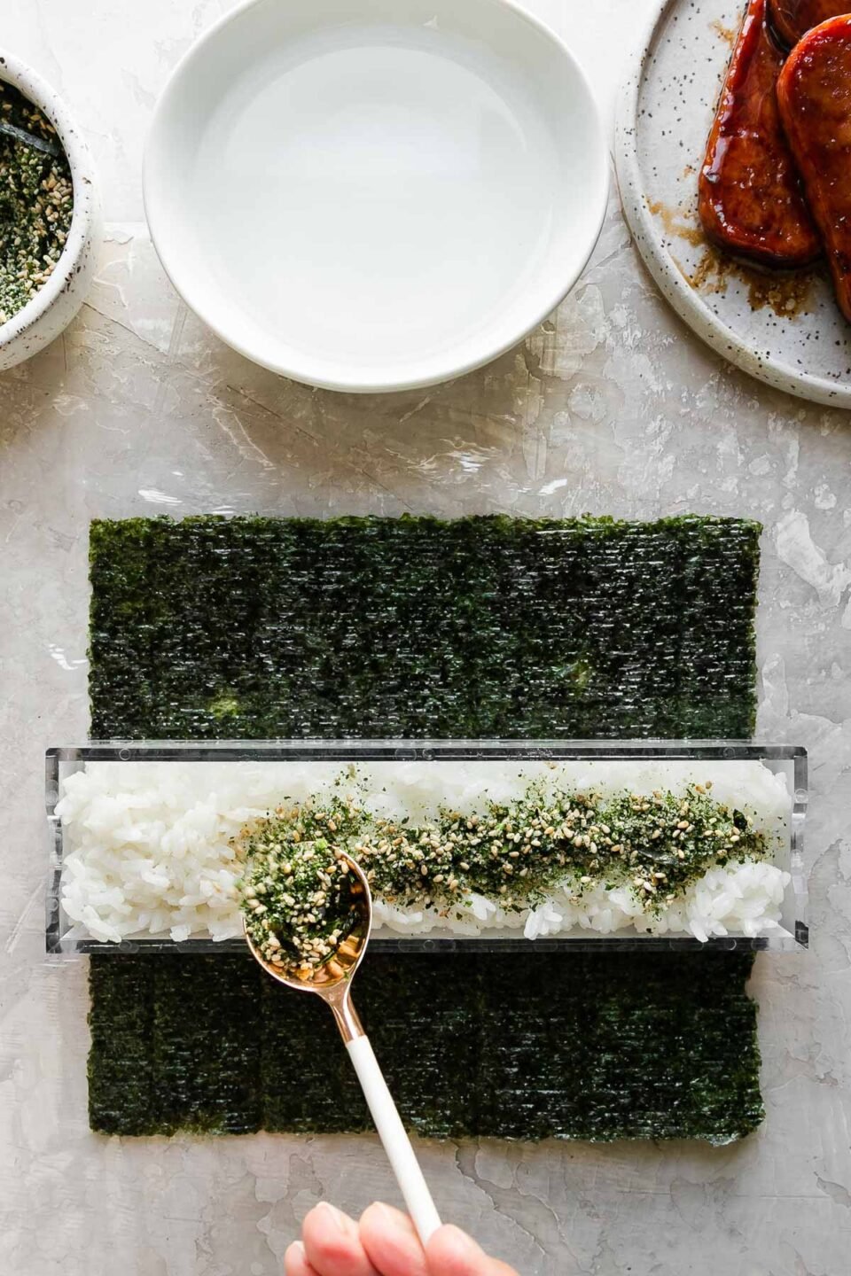 A close up of a woman's hand holding a white and gold spoon to sprinkle a generous amount of furikake seasoning atop a rice filled outer box of a double musubi mold. The musubi mold rests atop a single sheet of sushi nori arranged on a piece of plastic wrap atop a creamy white textured surface. Resting above the nori & the musubi mold is a small bowl filled with furikake seasoning with a spoon resting inside, another small white bowl filled with water, and a small speckled ceramic plate with pan-fried teriyaki Spam.