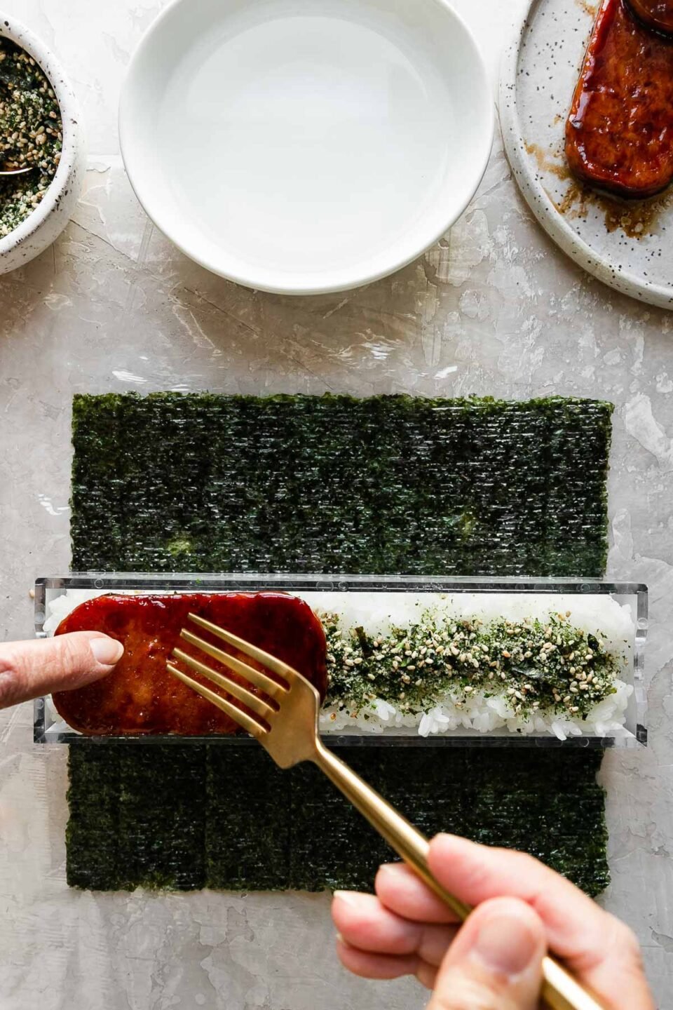 A close up of a woman using her hands and a fork to top a rice filled and furikake seasoned outer box of a double musubi mold with a slice of pan-fried teriyaki Spam. The musubi mold rests atop a single sheet of sushi nori arranged on a piece of plastic wrap atop a creamy white textured surface. Resting above the nori & the musubi mold is a small bowl filled with furikake seasoning with a spoon resting inside, another small white bowl filled with water, and a small speckled ceramic plate with pan-fried teriyaki Spam.