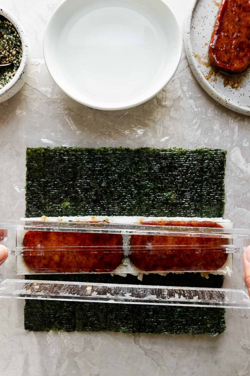 A close up of a woman using her hands to remove the lid of a spam musubi filled musubi mold to create a finished spam musubi. Resting above the musubi mold is a small bowl filled with furikake seasoning with a spoon resting inside, another small white bowl filled with water, and a small speckled ceramic plate with pan-fried teriyaki Spam.