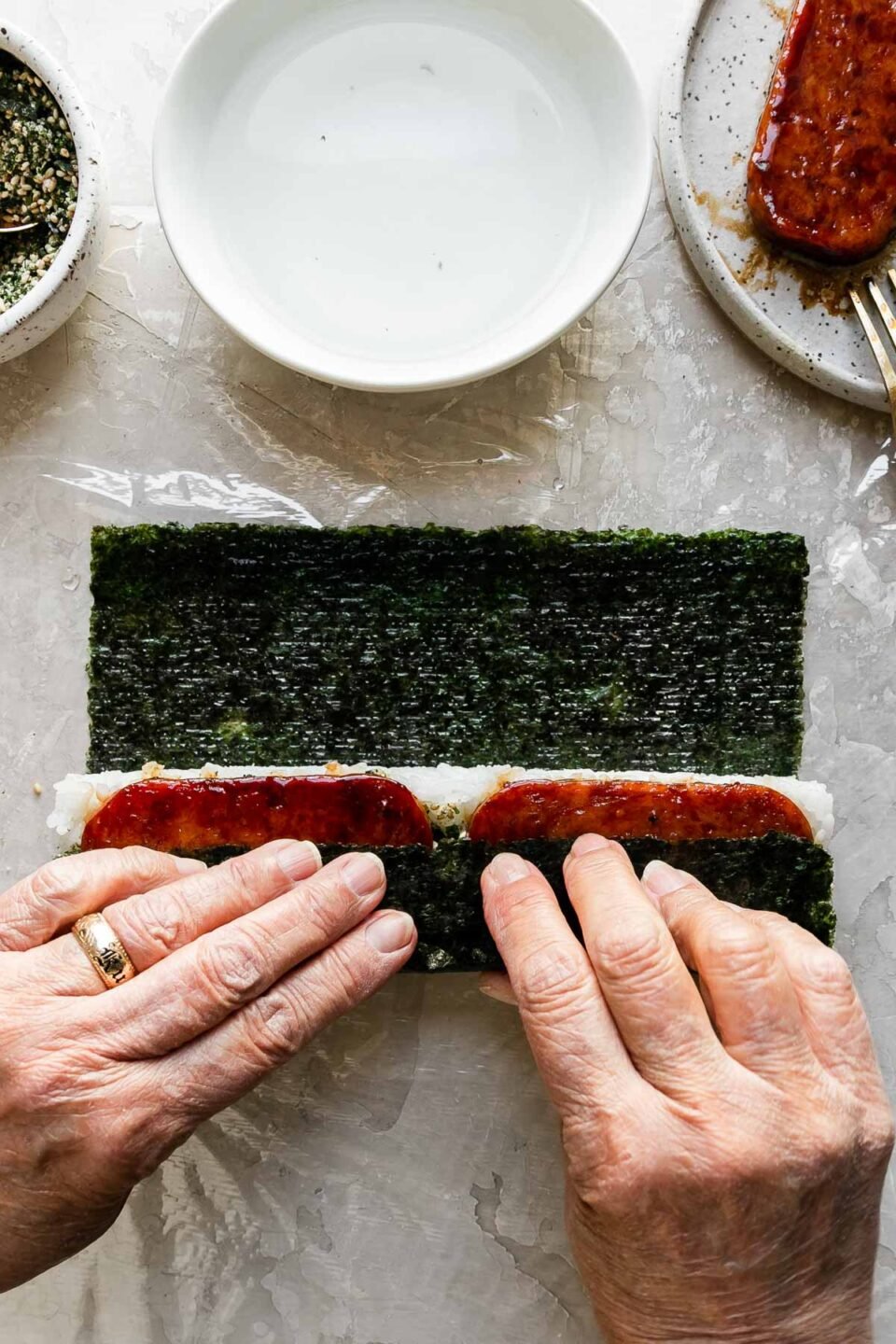 A close up of a woman's hands work to pull the side of the nori sheet closest to her up and over the inside of the spam musubi. Resting above the almost finished musubi is a small bowl filled with furikake seasoning with a spoon resting inside, another small white bowl filled with water, and a small speckled ceramic plate with pan-fried teriyaki Spam.