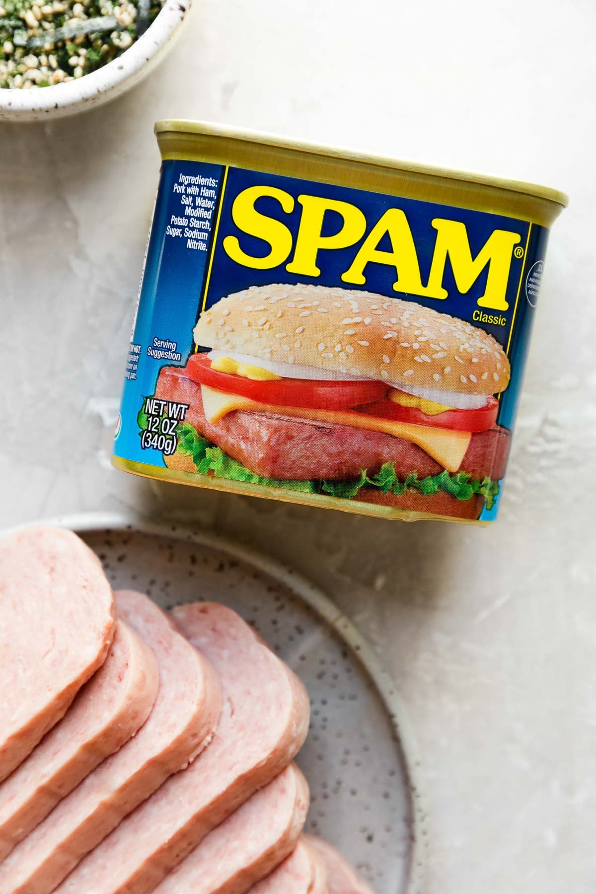 A can of Spam lays flat with the label facing upwards on a creamy white textured surface. A small ceramic plate topped with sliced pieces of spam rests below the can while a small white speckled ceramic bowl filled with furikake seasoning sits above it.