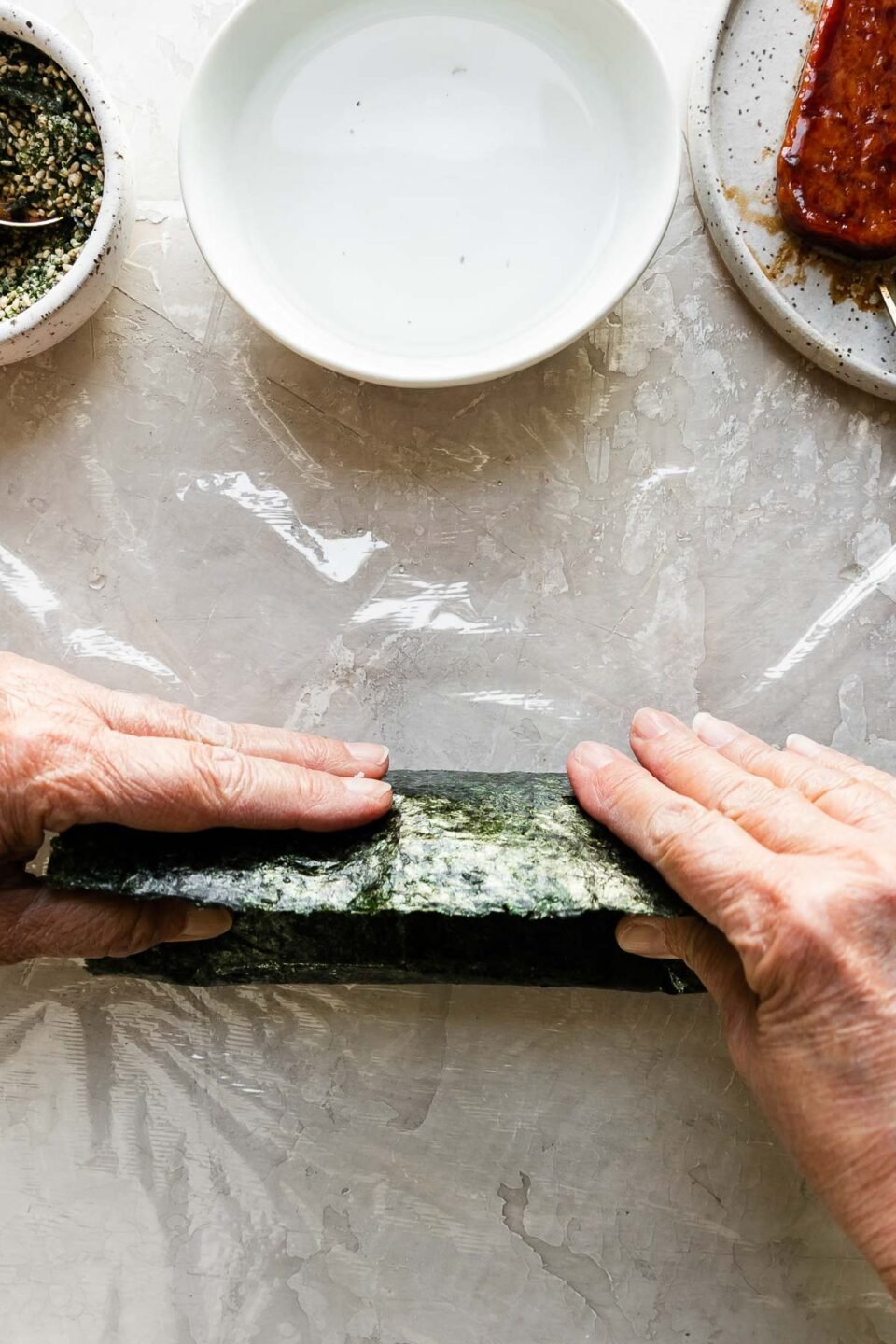 A close up of a woman's hands working to roll a partially wrapped musubi away from herself until it is sealed shut. Positioned just above the almost finished musubi is a small bowl filled with furikake seasoning with a spoon resting inside, another small white bowl filled with water, and a small speckled ceramic plate with pan-fried teriyaki Spam.