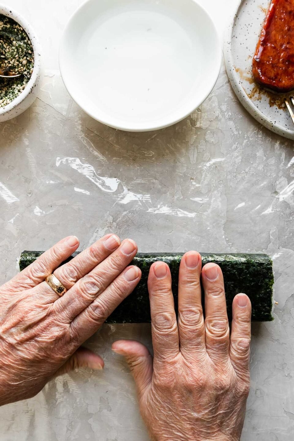 A close up of a woman's hands working to roll a partially wrapped musubi away from herself until it is sealed shut. Positioned just above the almost finished musubi is a small bowl filled with furikake seasoning with a spoon resting inside, another small white bowl filled with water, and a small speckled ceramic plate with pan-fried teriyaki Spam.