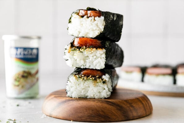 A stack of three Hawaiian spam musubi rest atop a wooden serving platter. The platter sits atop a creamy white textured surface. A container of Furikake seasoning and a plate of additional finished spam musubi sit out of focus in the background.