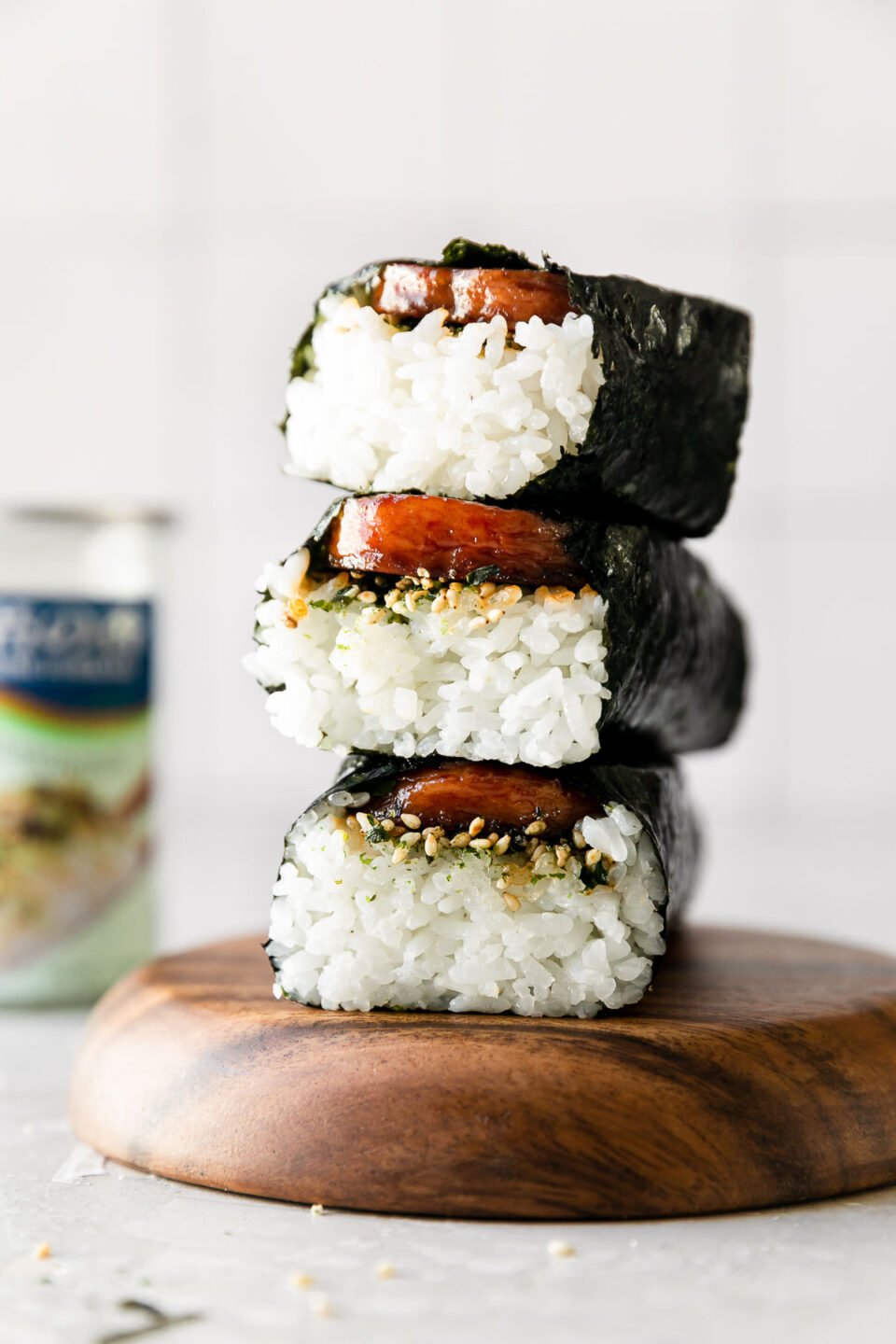 A stack of three Hawaiian spam musubi rest atop a wooden serving platter. The platter sits atop a creamy white textured surface and a container of Furikake seasoning sits out of focus in the background.
