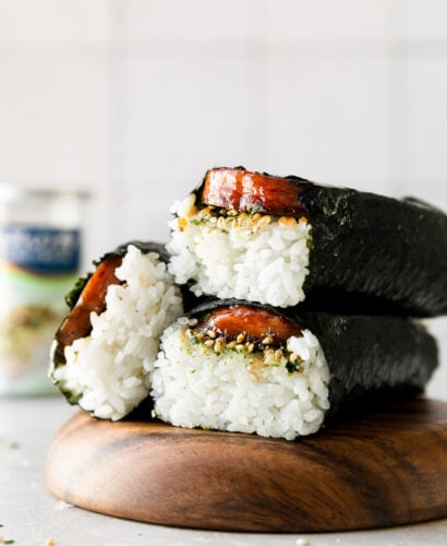 Three Hawaiian spam musubi are arranged atop a wooden serving platter. The platter sits atop a creamy white textured surface and a container of Furikake seasoning sits out of focus in the background.