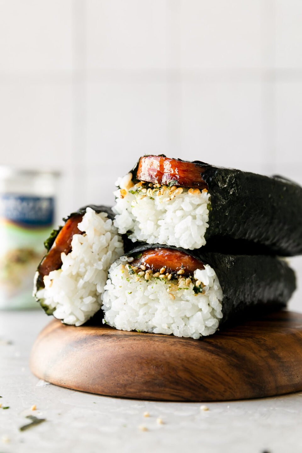 Three Hawaiian spam musubi are arranged atop a wooden serving platter. The platter sits atop a creamy white textured surface and a container of Furikake seasoning sits out of focus in the background.