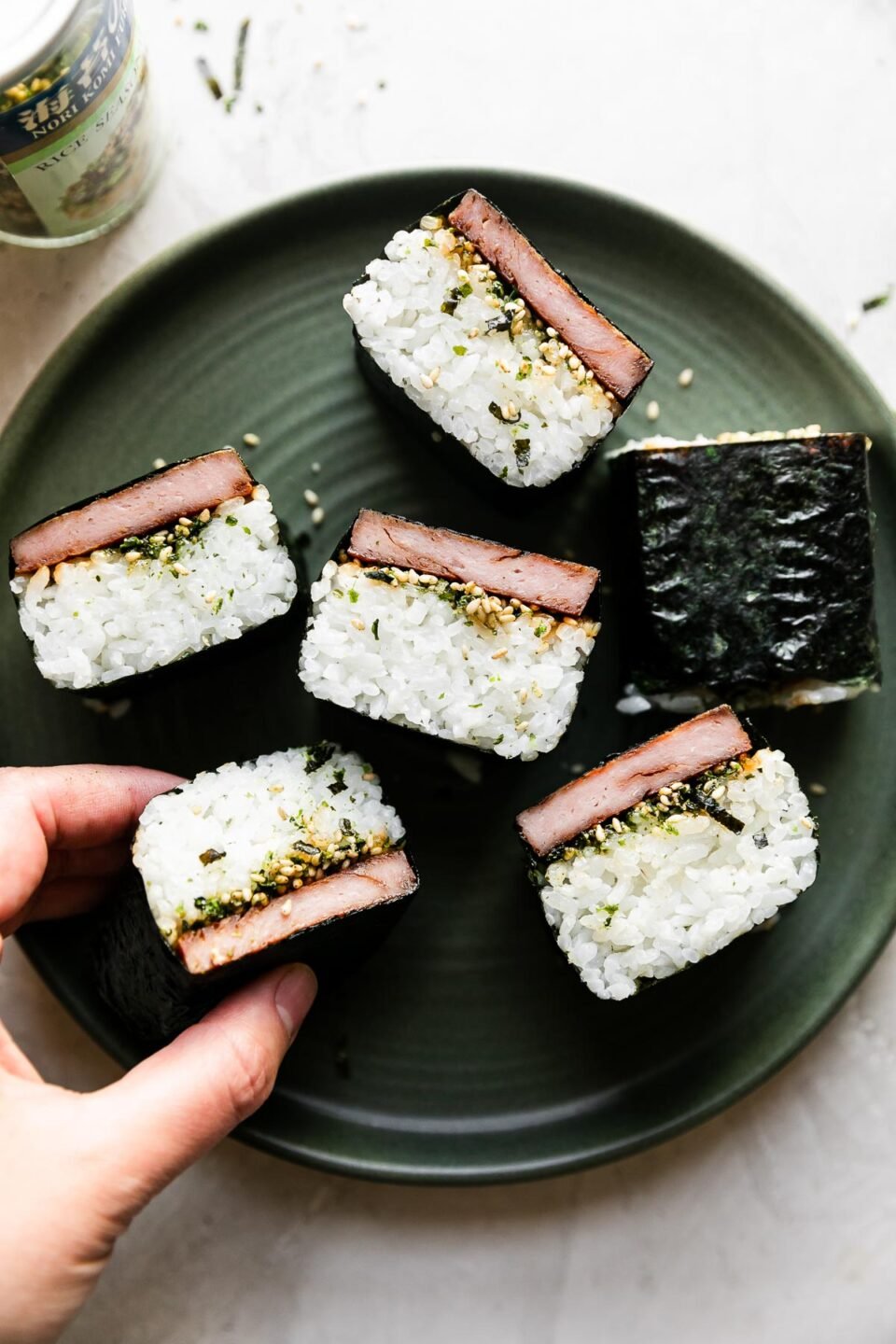 Six finished spam musubi are arranged atop a green ceramic plate. The plate sits atop a creamy white textured surface. A container of Furikake seasoning rests alongside the plate, while a woman's hand reaches for one of the musubi.