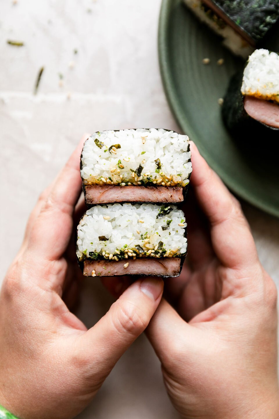 A woman's hands hold two stacked Hawaiian spam musubi above a creamy white textured surface. A green ceramic plate with additional musubi sits atop the creamy white textured surface slightly out of focus.