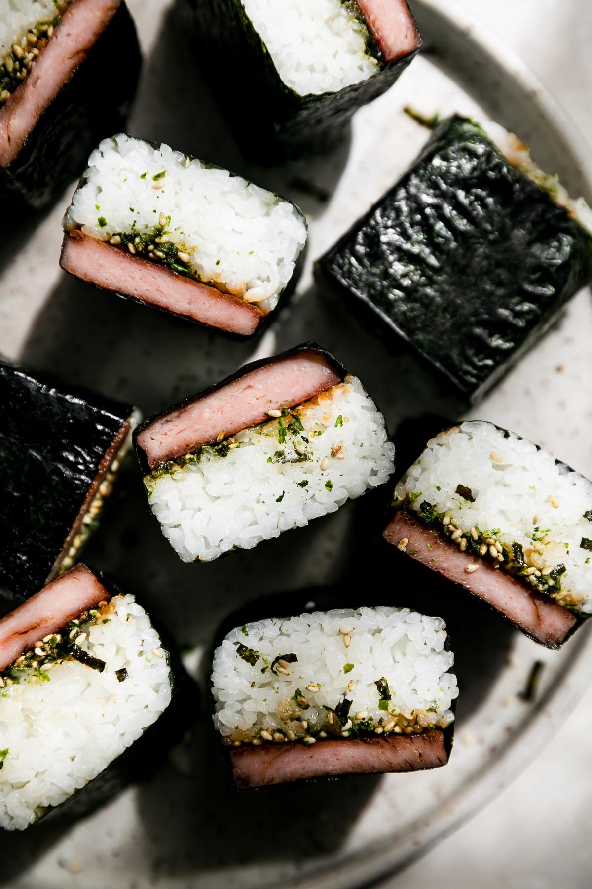 Nine finished spam musubi sit atop a gray speckled ceramic plate atop a creamy white textured surface.