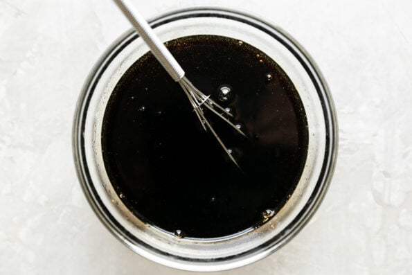 Prepared teriyaki sauce for spam musubi fills a clear glass mixing bowl that sits atop a creamy white textured surface. A small wire whisk rests inside of the teriyaki sauce for mixing.