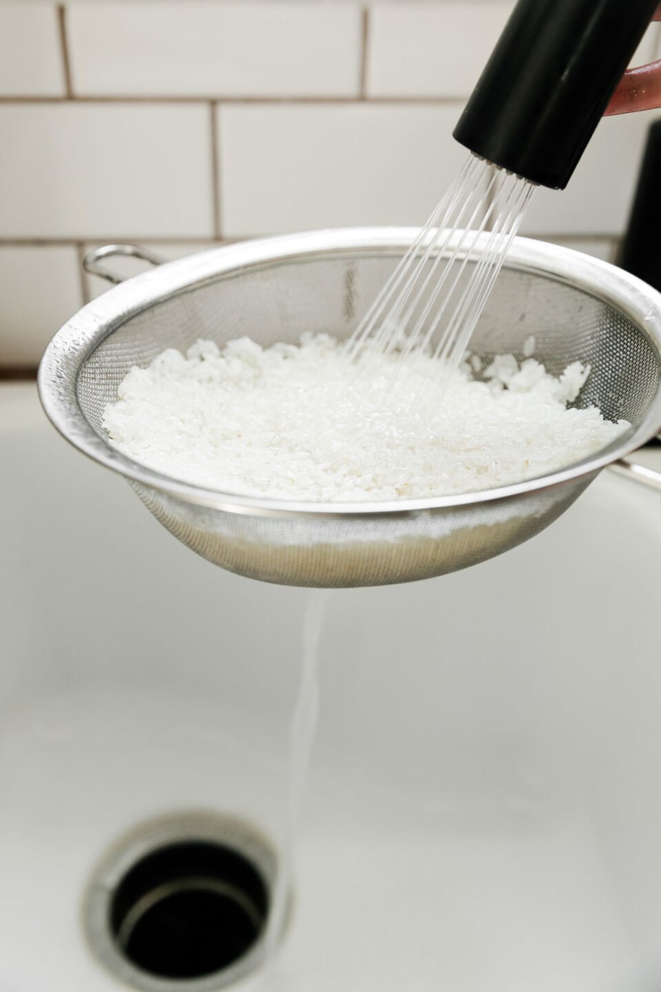 White rice fills a fine mesh colander while being rinsed well underneath a stream of water from a faucet as it is held over a sink. The water drains into the sink below. A white subway tile is seen in the background.