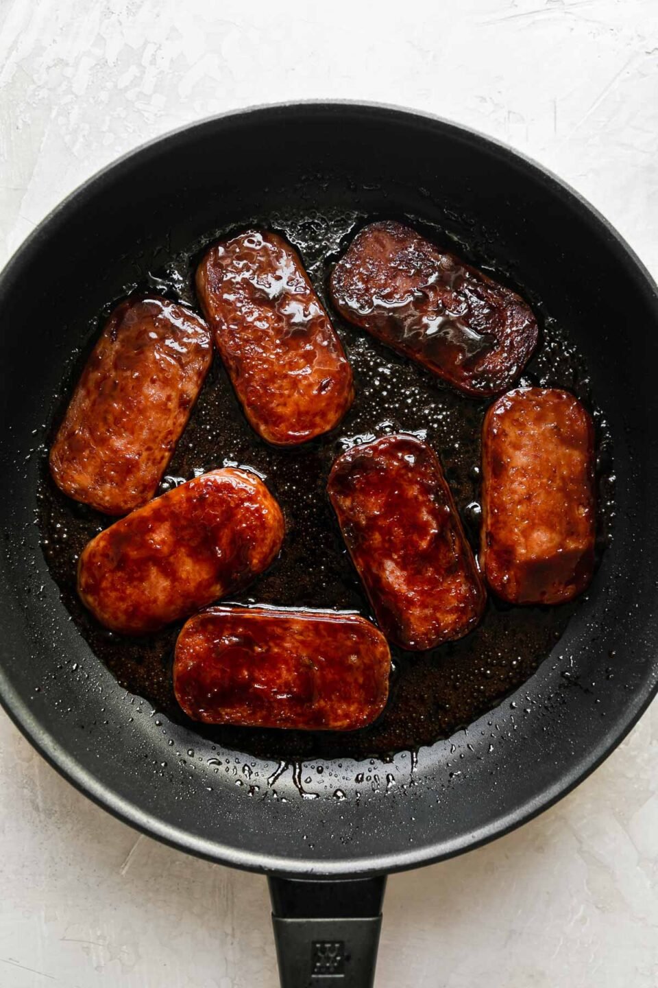 Spam glazed with teriyaki sauce pan fries in a large black skillet. The pan sits atop a creamy white textured surface.