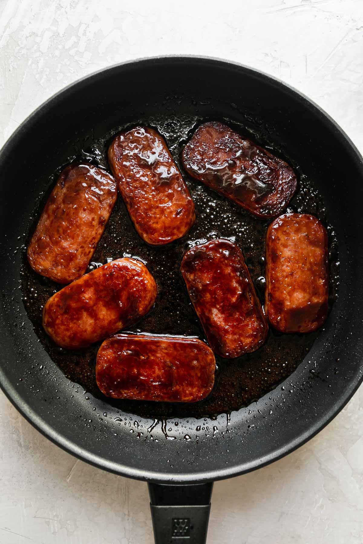 How to make Hawaiian spam musubi, step 3: Pan-fry the Spam & glaze with teriyaki sauce. Seven pieces of pan-fried spam that have been glazed with a homemade teriyaki sauce fill a black non-stick frying pan. The pan sits atop a creamy white textured surface.
