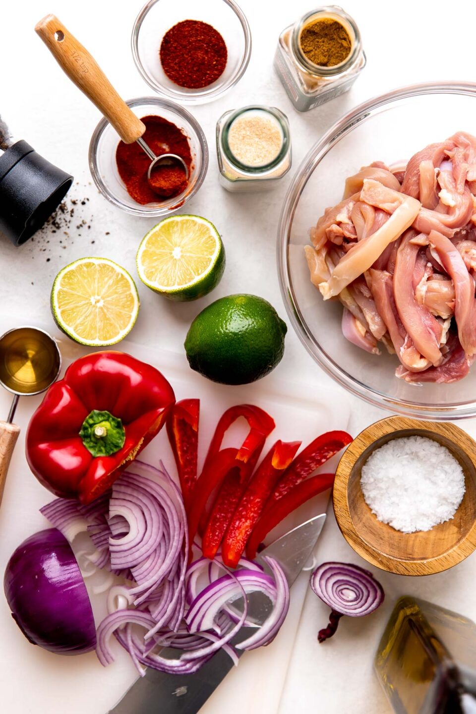 Skillet chicken fajita ingredients arranged on a creamy white textured surface: boneless, skinless chicken thighs or breasts, limes, olive oil, agave nectar, ancho chili powder, ground cumin, smoked paprika, garlic powder, bell pepper, red onion, kosher salt, and ground black pepper.