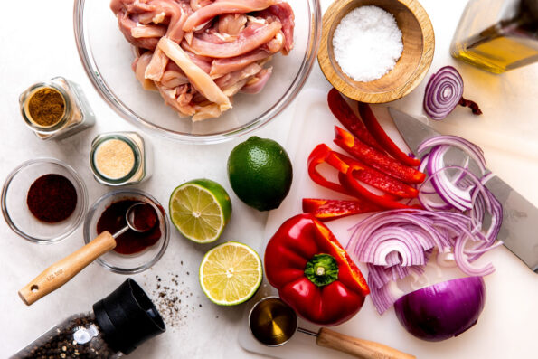 Skillet chicken fajita ingredients arranged on a creamy white textured surface: boneless, skinless chicken thighs or breasts, limes, olive oil, agave nectar, ancho chili powder, ground cumin, smoked paprika, garlic powder, bell pepper, red onion, kosher salt, and ground black pepper.