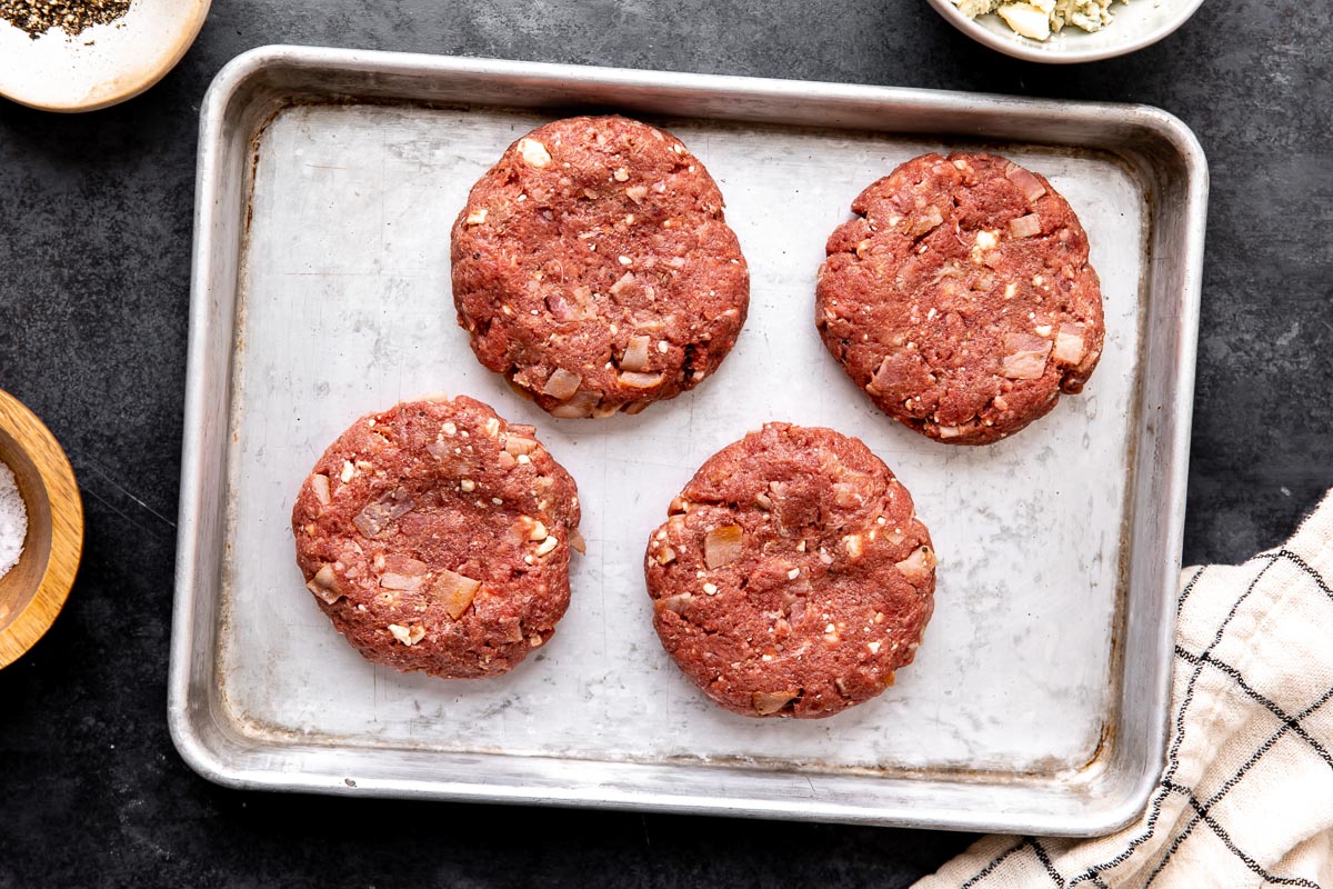 Four blue cheese burgers patties are arranged on a small aluminum baking sheet. The baking sheet sits atop a dark gray textured surface surrounded by a small white ceramic bowl filled with ground black pepper, a small wooden pinch bowl filled with kosher salt, a white and black window-pane patterned linen napkin, and a small white ceramic bowl filled with blue cheese crumbles.