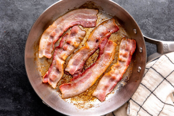 How to make blue cheese burgers, step 2: Par-cook the bacon. Five pieces of center-cut bacon par-cook in a stainless steel frying pan. The frying pan rests atop a dark gray textured surface with a black and white window-pane patterned linen napkin placed alongside.