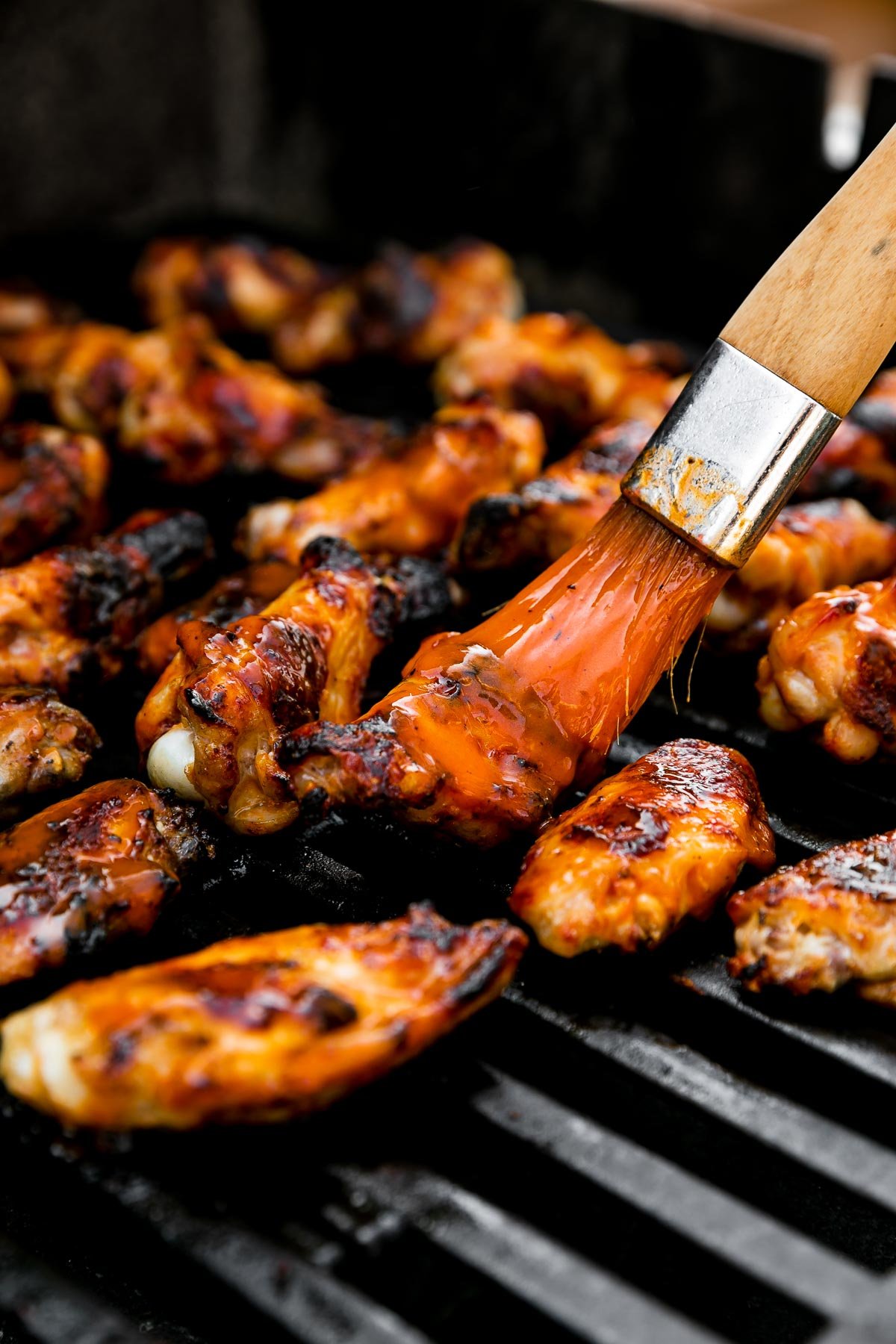 Grilled buffalo wings arranged on a gas grill over direct heat to char grill them. A pastry brush brushes additional Frank's RedHot over the wings.