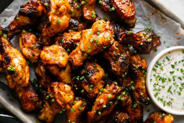 A close up of grilled buffalo wings served on a parchment lined aluminum baking sheet atop a creamy white textured surface. The wings have been garnished with freshly snipped chives and are served with a small bowl of ranch. A black linen napkin with white stripes rests alongside the baking sheet.