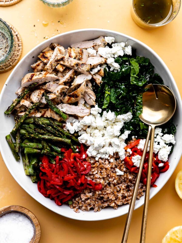 Finished grilled chicken and asparagus salad fills a white serving bowl with two gold serving spoons resting inside. The bowl is surrounded by a small wooden pinch bowl filled with kosher salt, lemon halves, a clear glass jar filled with lemon thyme vinaigrette, two clear glasses, and two rattan coasters that sit atop a yellow surface.