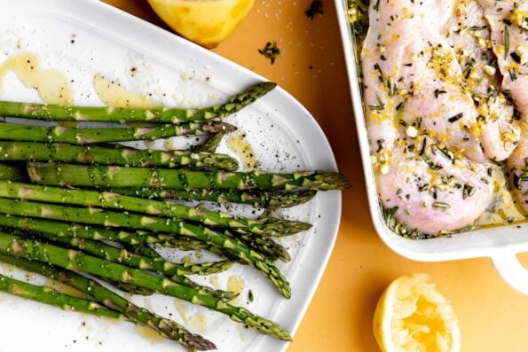 How to make grilled chicken and asparagus salad, step 5: Grill the chicken and asparagus. Marinated chicken and seasoned asparagus rest inside a white baking dish and atop a white serving platter respectively. The dishes sit atop a yellow surface with lemon halves surrounding.