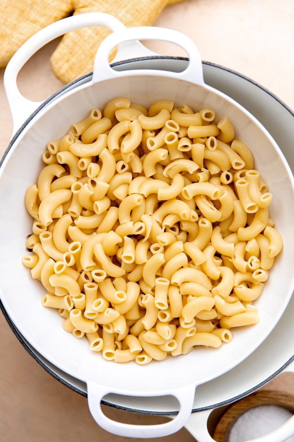 A white colander filled with al dente macaroni pasta for hawaiian macaroni salad rests inside of a large white double handle pot to drain. The pot sits atop a peach colored textured surface and is surrounded by a creamy yellow colored oven mitt and a small wooden pinch bowl filled with kosher salt.