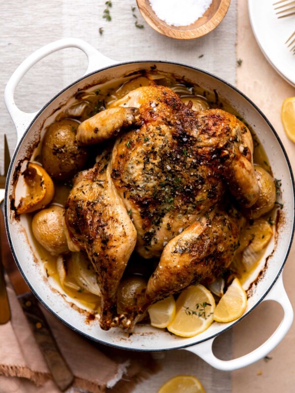 How to make spatchcock chicken over potatoes, step 5: Roast the spatchcock chicken. A finished roasted spatchcock chicken rests inside of a white double-handle oven-safe skillet. The skillet sits atop a cream colored textured surface with neutral colored linens resting underneath the skillet. A small wooden pinch bowl filled with kosher salt, lemon wedges, a carving fork, and two white plates with gold silverware surround the skillet at center.