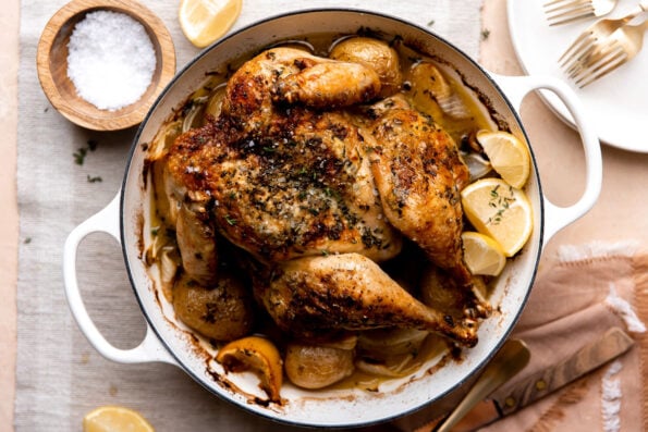 A finished roasted spatchcock chicken over potatoes rests inside of a white double-handle oven-safe skillet. The skillet sits atop a cream colored textured surface with neutral colored linens resting underneath the skillet. A small wooden pinch bowl filled with kosher salt, lemon wedges, a carving fork, and two white plates with gold silverware surround the skillet at center.