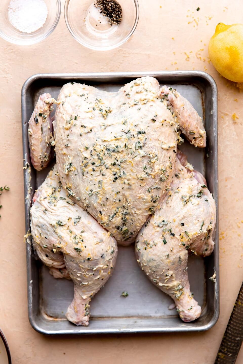 How to make lemon spatchcock chicken, step 3: Butter the spatchcock chicken. A spatchcock chicken lays flat atop an aluminum baking sheet that sits atop a cream colored textured surface. The spatchcock chicken has been covered in homemade lemon herb butter. Two clear glass pinch bowls are filled with kosher salt & ground black pepper, a lemon that has been zested, and a microplane surround the bowl at center.