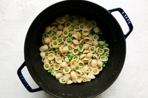 How to make lamb pasta, step 3: Boil the pasta. Cooked orecchiette pasta and shelled English peas fill a blue Staub cocotte that sits atop a creamy white textured surface.