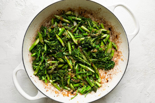 How to make lamb pasta, step 5: Cook the aromatics & spring veggies. Yellow onion, garlic, asparagus and broccolini cook inside of a white double-handle braiser. The braiser sits atop a creamy white textured surface.