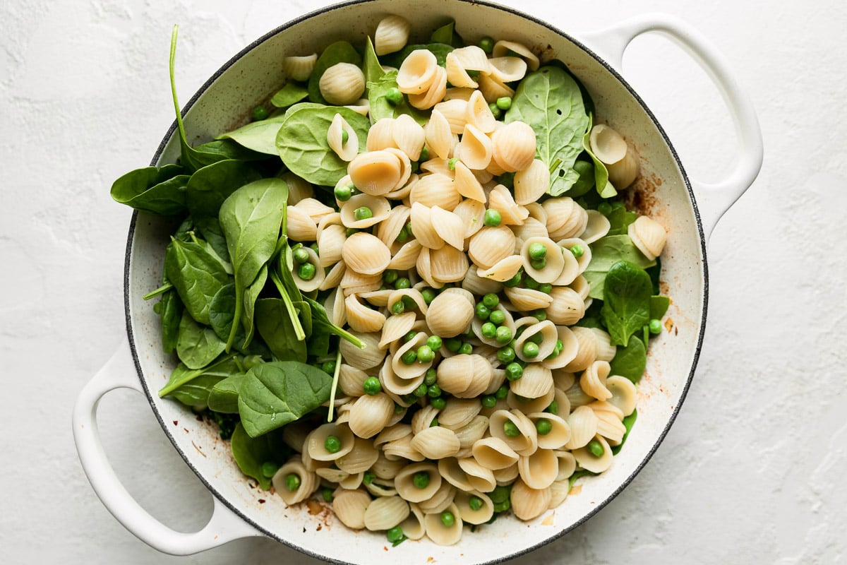 How to make lamb pesto pasta, step 6: Build the pasta. Baby spinach and cooked pasta and peas are added atop cooked aromatics inside of a white double-handle braiser. The braiser sits atop a creamy white textured surface.