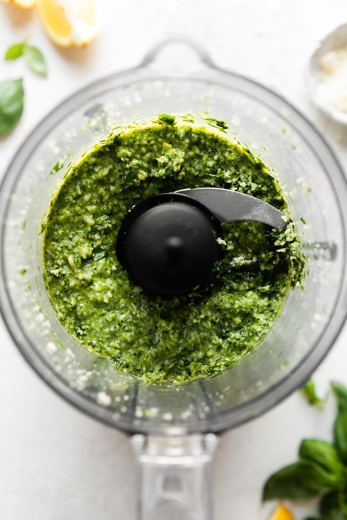 Blended lemon basil pesto in a food processor carafe, atop a white surface surrounded by additional pesto ingredients that are out of focus.